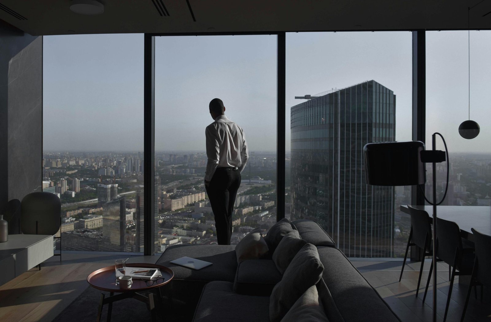 A man in business attire gazing out of a luxury apartment’s floor-to-ceiling window, overlooking a city skyline.